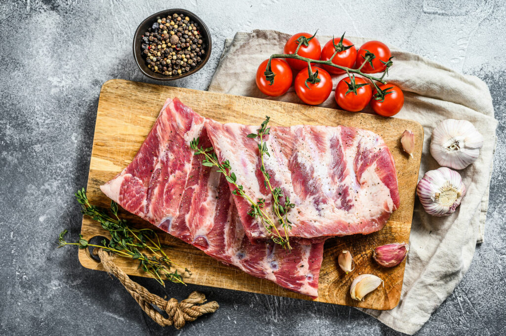 Raw pork with herbs on a wooden cutting board surrounded by tomatos, black peppers and garlic on a greystone background